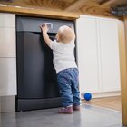 A one year old child is standing in the kitchen with dishwasher and is pressing buttons on the control panel of the dishwasher. He is wearing blue trousers and a bright shirt. A navy ball lies on the floor.
