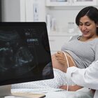 Kobieta w ciąży leży na łóżku w gabinecie lekarskim i podpiera się łokciami. Patrzy na swój brzuch. Lekarz przeprowadza USG płodu. Widać obraz płodu na monitorze. 