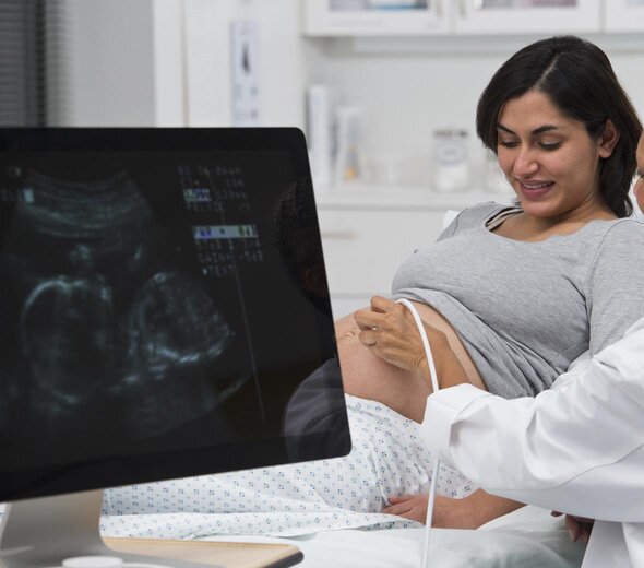 Kobieta w ciąży leży na łóżku w gabinecie lekarskim i podpiera się łokciami. Patrzy na swój brzuch. Lekarz przeprowadza USG płodu. Widać obraz płodu na monitorze. 