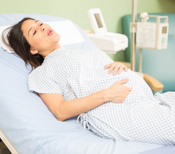 The expectant mother lies on a hospital bed and is dressed in hospital pajamas. The expectant mother is breathing quickly and deeply to ease pain. Mom supports her big tummy with both hands.