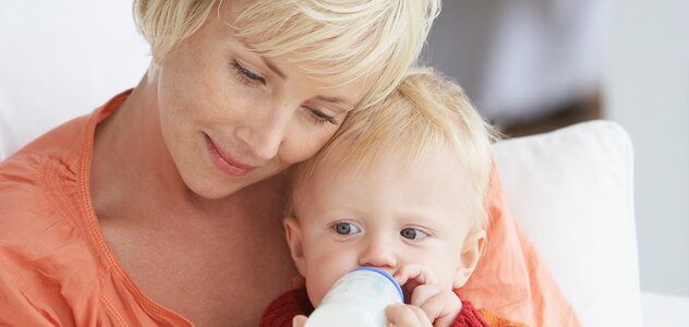 Mum sitting and holding on her lap a several months child who is drinking milk from a feeding bottle. The woman sitting behind the boy hugging her head to the baby's head.