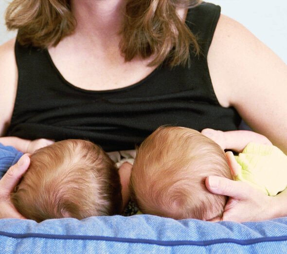 The mother visible in the picture supports two heads of twins with her hands. The babies are laid on their knees and facing the woman who puts them on her breast and feeds her mother's milk.