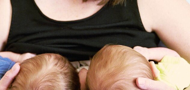The mother visible in the picture supports two heads of twins with her hands. The babies are laid on their knees and facing the woman who puts them on her breast and feeds her mother's milk.