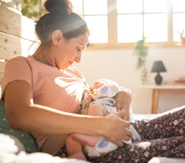 Mama siedzi na podłodze oparta o drewnianą szafkę. Karmi niemowlę piersią.  Ma długie brązowe włosy upięte w kok i różową bluzkę. Patrzy na dziecko. Uśmiecha się. W oddali widać okno, przez które wpadają promienie słoneczne. 