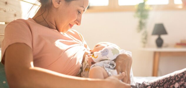 Mama siedzi na podłodze oparta o drewnianą szafkę. Karmi niemowlę piersią.  Ma długie brązowe włosy upięte w kok i różową bluzkę. Patrzy na dziecko. Uśmiecha się. W oddali widać okno, przez które wpadają promienie słoneczne. 