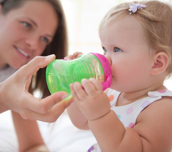 Mom holds the child a cup of non-spill to the mouth. The girl holds it with hands and drinks a drink  independently, woman lying next to a child on the bed.