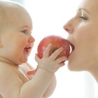 Dziecko jest naprzeciwko mamy, jest nago. Trzyma w rączce jabłko. Mam gryzie jabłko. Mama ma blond włosy. 