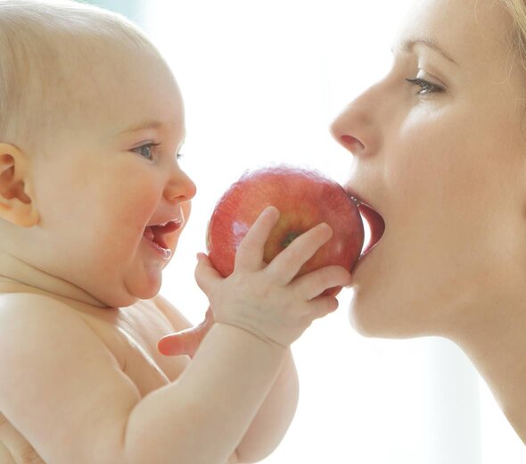 Dziecko jest naprzeciwko mamy, jest nago. Trzyma w rączce jabłko. Mam gryzie jabłko. Mama ma blond włosy. 