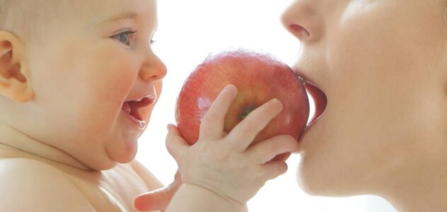 Dziecko jest naprzeciwko mamy, jest nago. Trzyma w rączce jabłko. Mam gryzie jabłko. Mama ma blond włosy. 