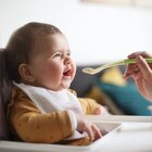 W krzeselku do karmienia siedzi dziecko. Ubrane jest w pomarańczową bluzeczkę w brązowe kropki. Pod brodą ma śliniak. Dziecko jest karmione łyżeczką.