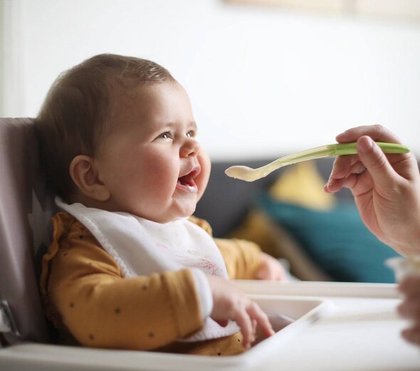 W krzeselku do karmienia siedzi dziecko. Ubrane jest w pomarańczową bluzeczkę w brązowe kropki. Pod brodą ma śliniak. Dziecko jest karmione łyżeczką.