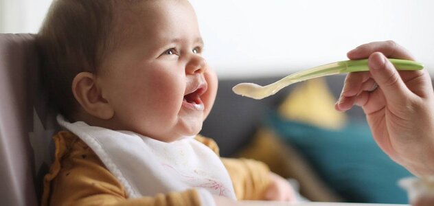 W krzeselku do karmienia siedzi dziecko. Ubrane jest w pomarańczową bluzeczkę w brązowe kropki. Pod brodą ma śliniak. Dziecko jest karmione łyżeczką.