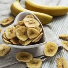 Na szarych podkładkach na stole stoi biała miseczka w kszłcie serduszka. W miseczce leżą chipsy bananowe. Chipsy bananowe są też rozsypane na stole. Obok leżą dwa świeże banany.