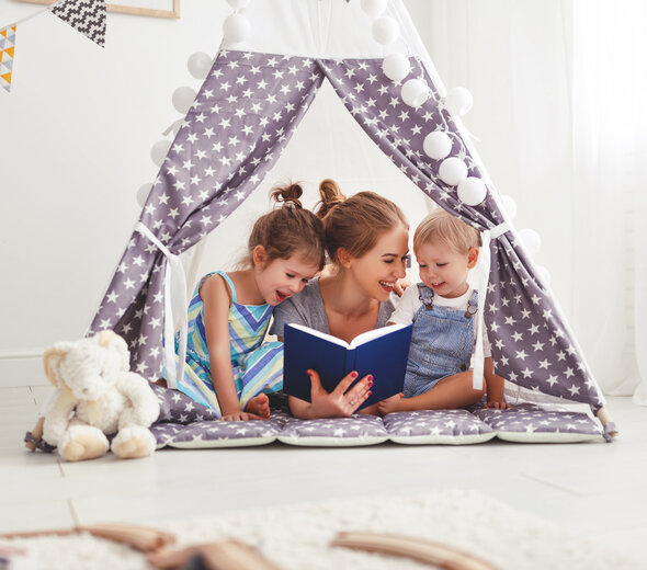 Mama i dwoje dzieci leżą na brzuchu w namiocie tipi ustawionym na podłodze w salonie. Tipie jest z fioletowego materiału w białe gwiazdki. Mama uśmiecha się do dzieci. Jedno dziecko jest starsze, drugie młodsze.  Mama ma spięte, długie blond włosy.  Trzyma w ręku granatową książkę. Obok siedzi biały królik przytulanka.