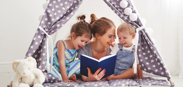 Mama i dwoje dzieci leżą na brzuchu w namiocie tipi ustawionym na podłodze w salonie. Tipie jest z fioletowego materiału w białe gwiazdki. Mama uśmiecha się do dzieci. Jedno dziecko jest starsze, drugie młodsze.  Mama ma spięte, długie blond włosy.  Trzyma w ręku granatową książkę. Obok siedzi biały królik przytulanka.