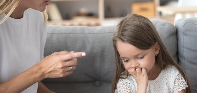 A young mother is sitting on a couch with her few-year-old daughter. Mom calmly but firmly explains to the girl what she did wrong by threatening her with her finger. Toddler looks down, supporting his chin with his hand.