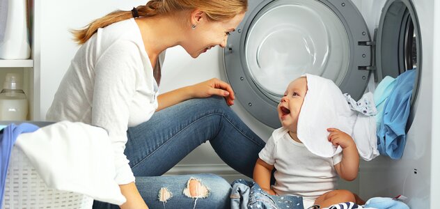 A young mother and her several-month-old baby are in the home laundry. The parent sits on the floor next to the sitting infant and the standing washing machine.