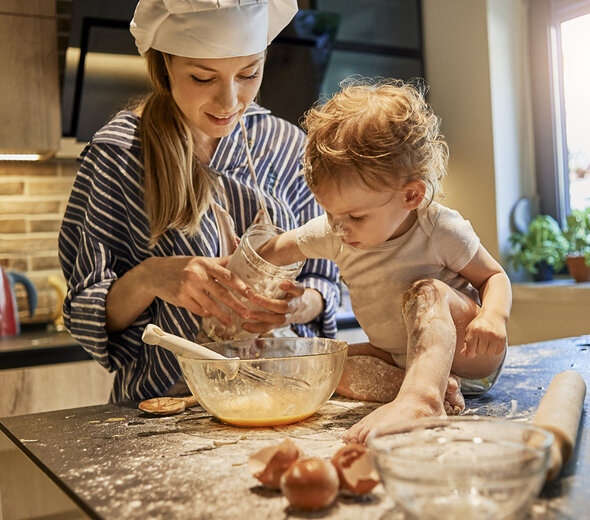 A young mother spends time with her several-month-old baby in the kitchen. A woman is making cookies with the baby. The infant is sitting on the counter alone and is helping mom in cooking.