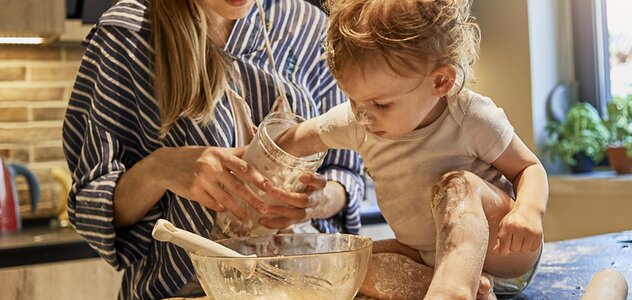 A young mother spends time with her several-month-old baby in the kitchen. A woman is making cookies with the baby. The infant is sitting on the counter alone and is helping mom in cooking.