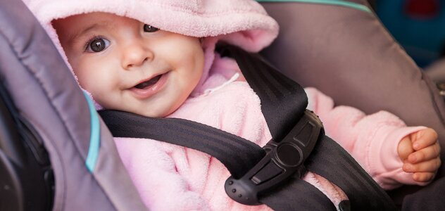 A recently born baby with a wide smile ia sitting calmly in a baby car seat. Baby is wearing in a pink sweatshirt with tiny ears sewn on the hood.