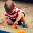 A dozen months old baby is outside, in the open air. The infant is playing in the sand by gouging in it with a blue spatula. The child is sitting alone on sand in the sandbox.