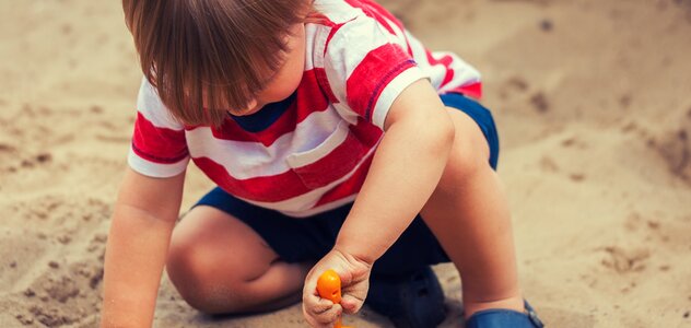 A dozen months old baby is outside, in the open air. The infant is playing in the sand by gouging in it with a blue spatula. The child is sitting alone on sand in the sandbox.