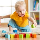 Mały chłopczyk siedzi na drewnianej podłodze. Ma blond włosy, niebieskie spodnie i żółtą koszulkę z krótkim rękawem. Ma bose stópki.