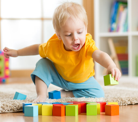 Mały chłopczyk siedzi na drewnianej podłodze. Ma blond włosy, niebieskie spodnie i żółtą koszulkę z krótkim rękawem. Ma bose stópki.