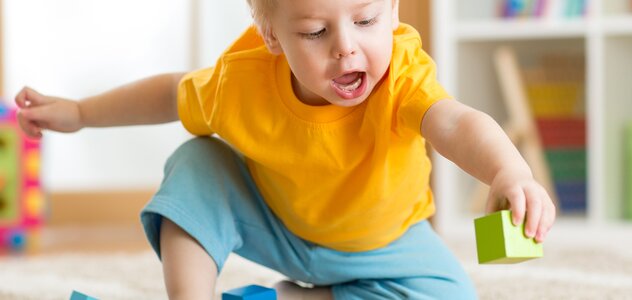 Mały chłopczyk siedzi na drewnianej podłodze. Ma blond włosy, niebieskie spodnie i żółtą koszulkę z krótkim rękawem. Ma bose stópki.