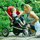 The boy sits in a red pram and holds a bitten apple in his hands. The mother squats next to the child and smiles at him. The walk takes place against the background of a green park.