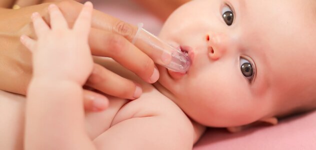 A recently born baby lying in the middle of a large bed. Baby is lying on his back on a pink sheet without clothes, only in a diaper. Mom trying to massage the baby's gums with a rubber brush on her finger.