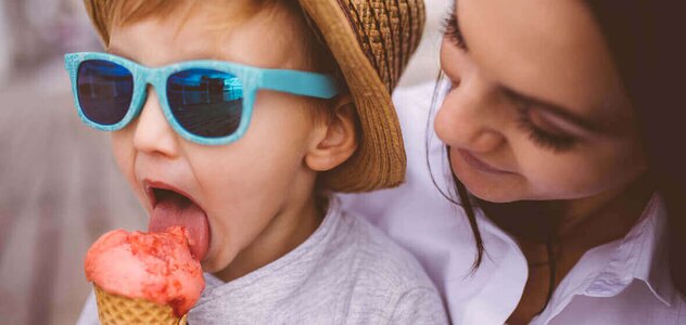   Poznaj wymowę A young mother enjoys spending time with her little son. The boy wearing blue glasses, a straw hat and a sweatshirt, he is sitting on his parent's knees and eats strawberry ice cream.