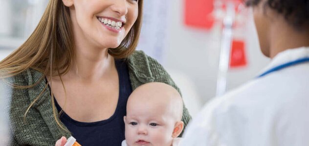 Uśmiechnięta kobieta trzyma na ręku niemowlę w białym body, w drugiej dłoni ma opakowanie z lekami. Naprzeciw stoi lekarka w kitlu, która udziela porady młodej matce.