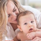 Niemowlę leży na brzuszku i patrzy w obiektyw. Ma ciemne blond włoski i brązowe oczy. Nie ma ubranka. Mama pochyla się nad niem przytula i całuję w tył główki. Ma długie blond falowane włosy i białą koszulę.