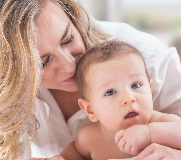 Niemowlę leży na brzuszku i patrzy w obiektyw. Ma ciemne blond włoski i brązowe oczy. Nie ma ubranka. Mama pochyla się nad niem przytula i całuję w tył główki. Ma długie blond falowane włosy i białą koszulę.