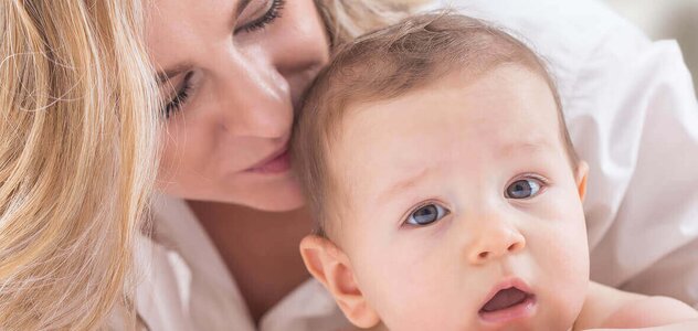 Niemowlę leży na brzuszku i patrzy w obiektyw. Ma ciemne blond włoski i brązowe oczy. Nie ma ubranka. Mama pochyla się nad niem przytula i całuję w tył główki. Ma długie blond falowane włosy i białą koszulę.